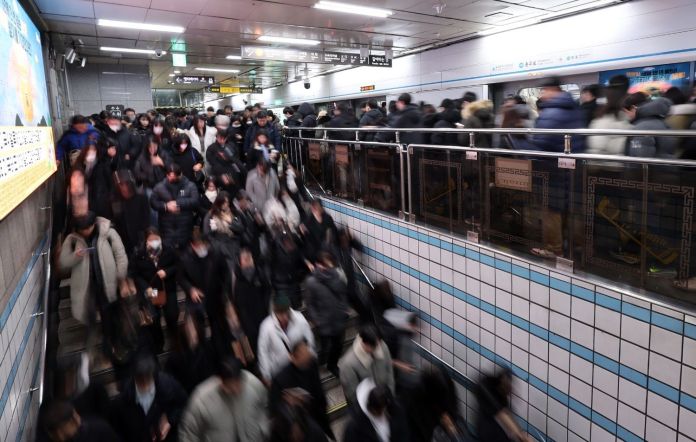 A crowded subway station with commuters walking down a staircase, many wearing masks and dark clothing, creating a blurred effect due to movement.