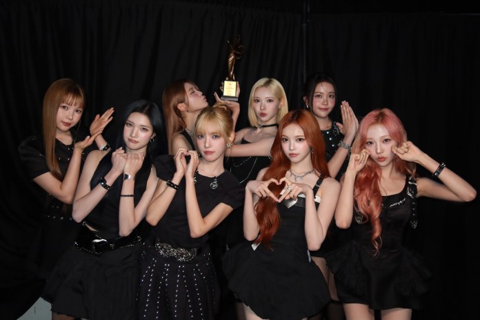 Hearts2Hearts members dressed in black outfits, posing in front of a trophy against a black backdrop.