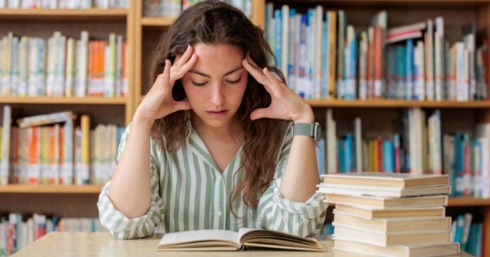 A young woman sitting at a table in a library, looking stressed while reading a book, with her hands on her head. Books are stacked beside her.