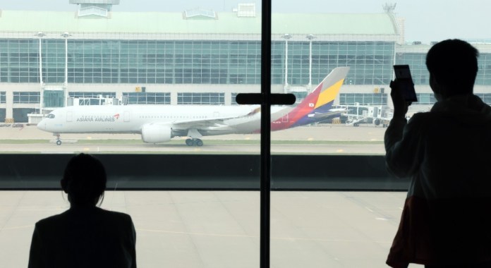 Silhouette of two people standing by a large window in an airport, one taking a photo, while an Asiana Airlines airplane taxis outside.