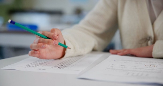 Close-up of a person's hand holding a green pencil while pointing at exam papers on a desk.