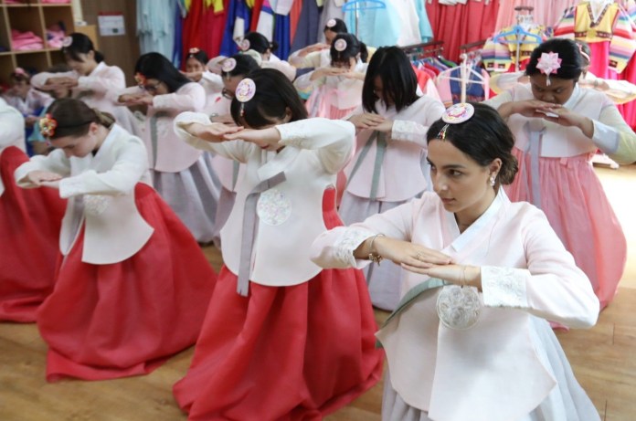 A group of women wearing traditional Korean hanbok outfits, practicing a dance routine in a dance studio with colorful clothing displayed in the background.