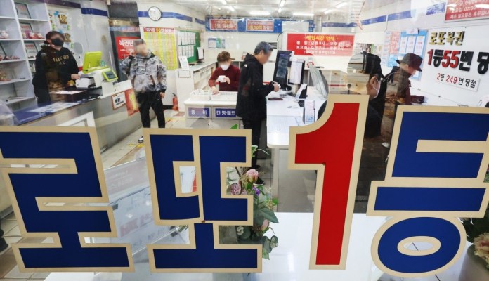 Interior view of a lottery ticket shop with several customers at the counter, displaying a large sign that reads '로또 1등' (Lotto 1st Prize) in bold letters.