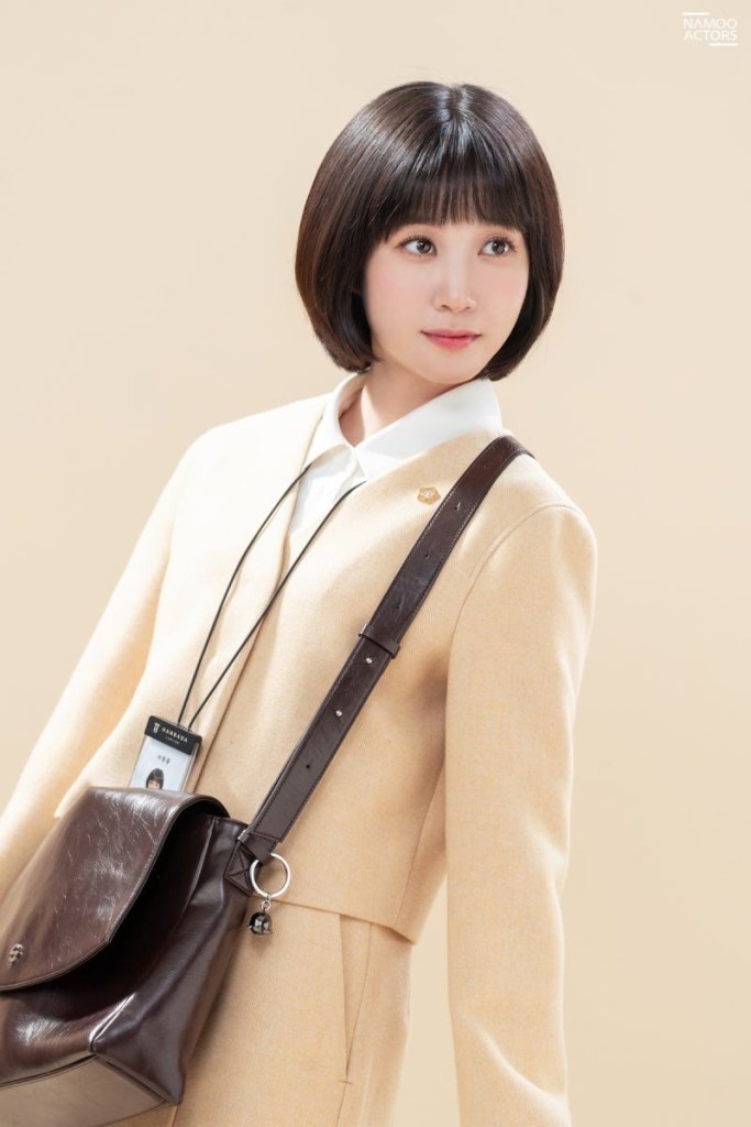 A young woman with a bob haircut, wearing a light beige suit and a white collared shirt, posing against a soft beige background. She has a brown shoulder bag and an ID badge hanging around her neck.