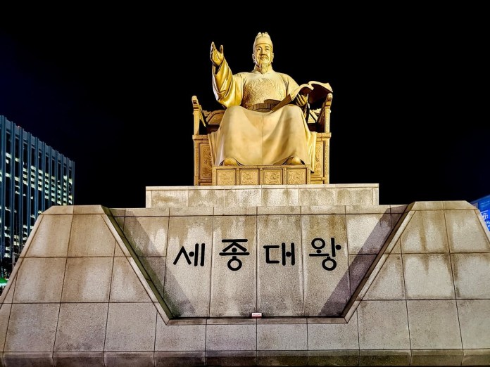 Statue of King Sejong the Great, illuminated at night, seated on a throne with a book in hand, with Korean text on the base.