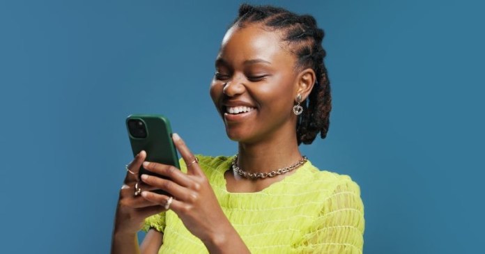 A woman with braided hair smiles while holding a green smartphone, wearing a bright yellow shirt.
