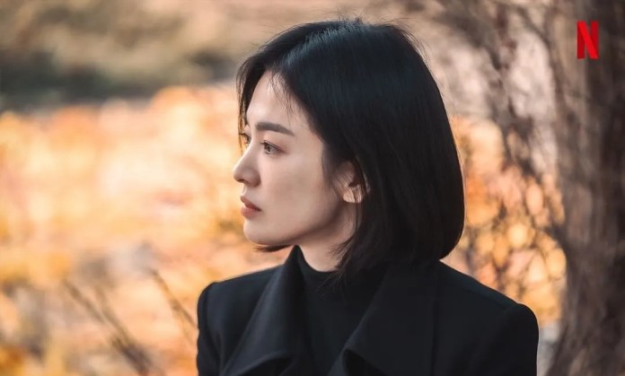 Side profile of a woman with short black hair, wearing a black coat, gazing thoughtfully into the distance with a blurred autumn background.