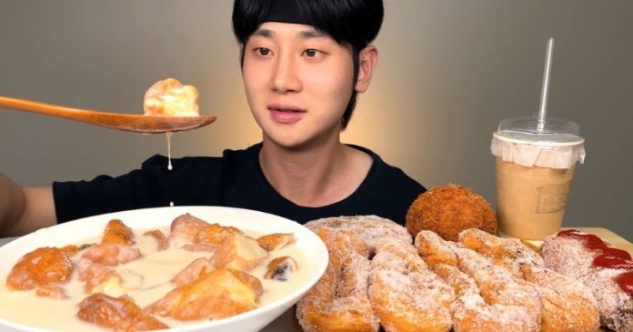 A person holding a spoonful of food, looking at a large bowl of dessert made with fried dough pieces and creamy sauce. A variety of fried snacks and a drink are arranged on the table.