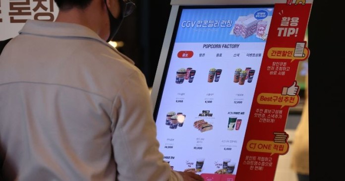 A person using a digital kiosk to view and order snack items at a movie theater, featuring various popcorn and beverage options and prices in Korean.