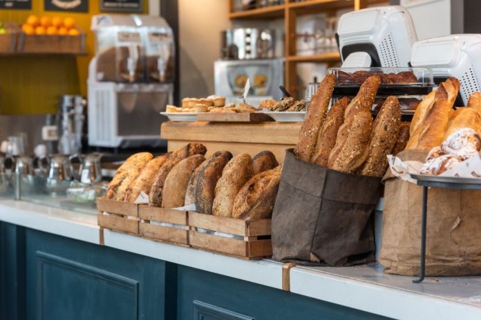 A display of various types of freshly baked bread, including baguettes and loaves, arranged in wicker baskets and brown paper bags at a cafe or bakery counter.