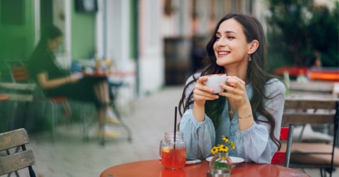 A woman sitting at a table in a café, smiling while holding a cup. A glass with a drink and a small vase with flowers are on the table, with another person in the background.