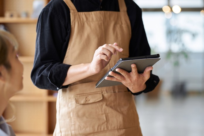 A barista in a brown apron is taking notes on a tablet while interacting with a customer.