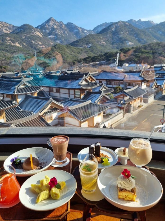 A beautiful outdoor view from a window showing traditional Korean houses with tiled roofs and mountains in the background. On the table, there are various delicious desserts and drinks, including a chocolate dessert, a creamy drink, a colorful cocktail, and a cake topped with a flower.