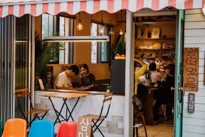 Interior view of a cozy cafe with a striped awning, two customers sitting at a table, and others socializing in the background.