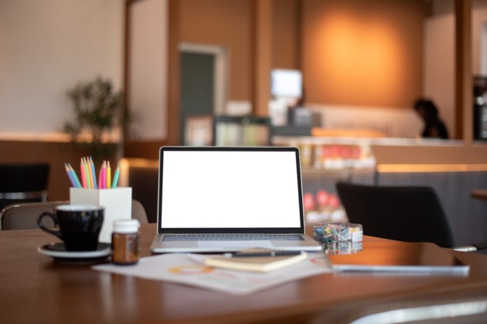A laptop with a blank screen on a wooden table in a cafe, accompanied by a coffee cup, a jar, colorful pens in a holder, and stationery items.