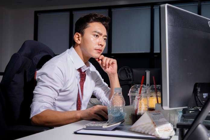 A focused young man in a white shirt and red tie sitting at a desk, working on a computer with several drinks nearby.