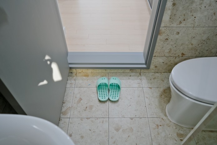 A pair of green bathroom slippers on tiled floor near a doorway leading to another room, with a toilet visible nearby.