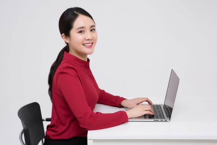 A young woman in a red blouse sits at a desk with a laptop, smiling while working.