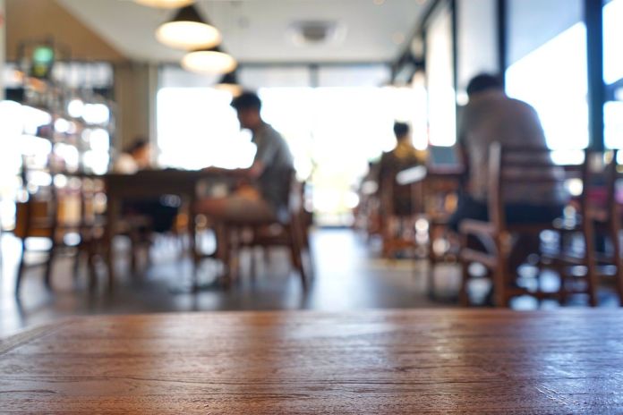 A blurred view of a busy coffee shop with wooden tables and people sitting at them, focusing on foreground wooden texture.