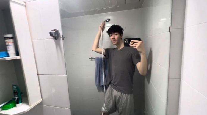 A person standing in a shower, holding a showerhead and taking a selfie. They are wearing a gray t-shirt and shorts, with a towel hanging nearby.