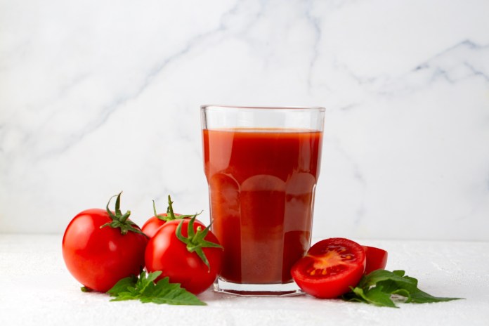A glass of tomato juice alongside fresh red tomatoes and a halved tomato, on a light marble background.