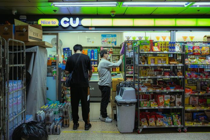 Two people inside a convenience store, with one person browsing shelves filled with snacks and drinks, and the other near the entrance. The store features a colorful signage with the words 'Nice to CU'.