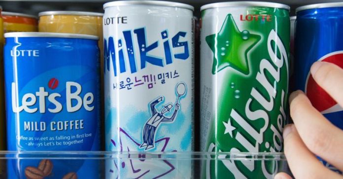A close-up of a beverage fridge featuring cans of Lotte drinks including Let's Be Mild Coffee, Milkis, and Chilsung Cider. A hand is seen reaching for a can.