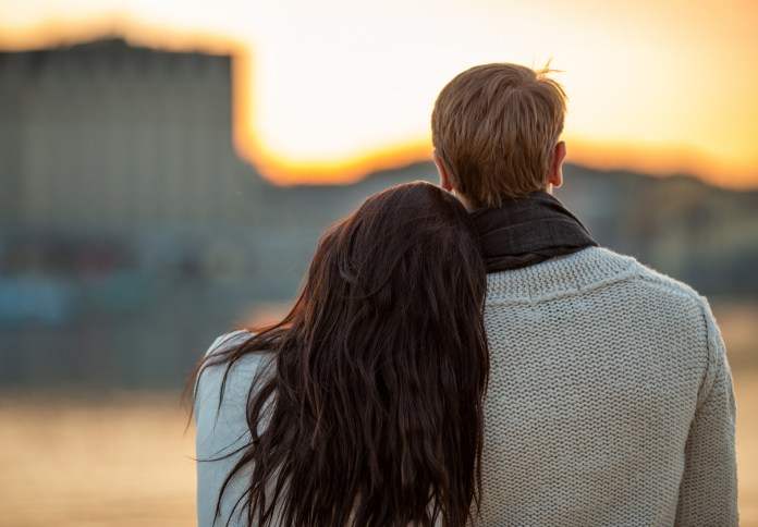 A couple embracing as they watch a sunset by the water, with the woman leaning her head on the man's shoulder.