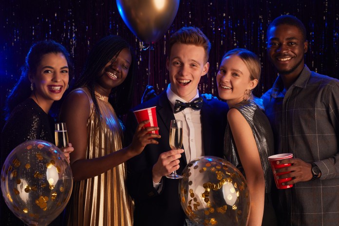 A group of five young adults celebrating at a party, smiling and holding drinks. They are dressed in festive attire with decorations in the background, including balloons and a shiny backdrop.