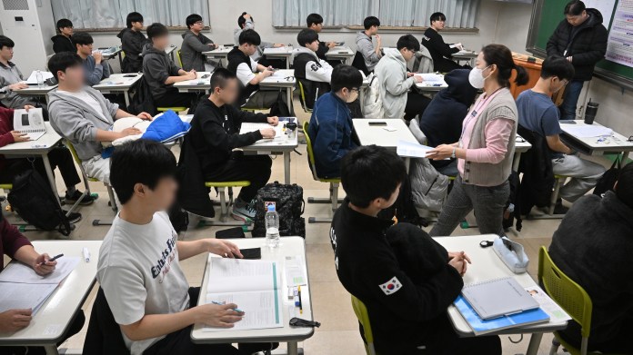A classroom filled with students seated at desks, focused on their studies while a teacher, wearing a mask, interacts with them.
