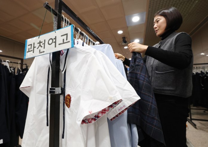 A woman organizing clothes on a rack in a clothing store, with a sign that reads '과천여고.' The clothing includes school uniforms with distinctive styles.