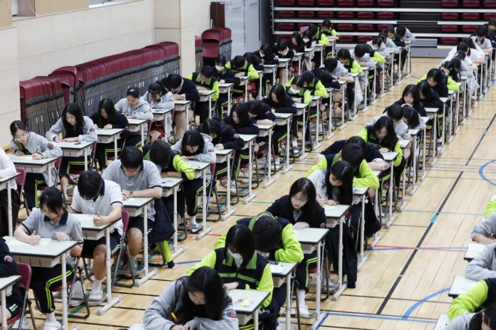Students seated at desks taking a test in a gymnasium, focused on their work.