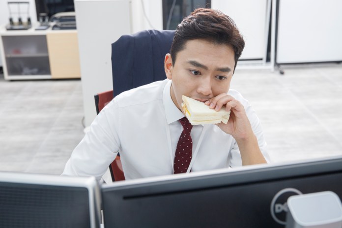A man in a white shirt and red tie sits at a desk with computers, holding a sandwich to his mouth while focused on his work.