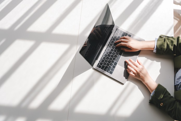 A person's hands typing on a laptop, positioned on a white table with shadows cast from nearby objects.