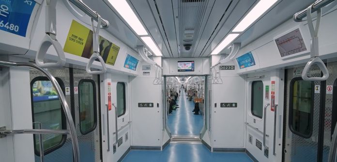Interior view of a modern subway train, featuring empty seating, grab handles, and digital display screens, with blue flooring and bright lighting.
