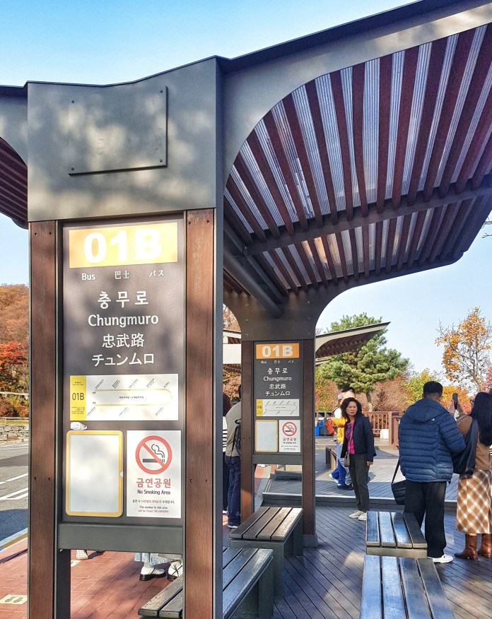 A bus stop labeled '01B Chungmuro' with bilingual signage in Korean and English. Several people are waiting and standing near the benches, surrounded by trees with autumn foliage.