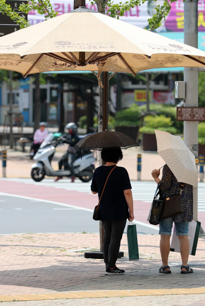 Two women standing under a large umbrella in a city street, one holding a shopping bag and the other holding a small umbrella. A motorcycle is visible in the background.
