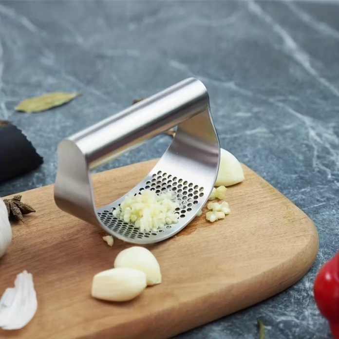 A stainless steel garlic press positioned on a wooden cutting board, with minced garlic and whole garlic cloves beside it.