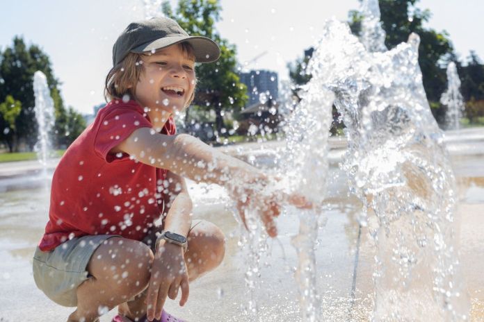 A young child in a red shirt and cap joyfully plays in a splash fountain, reaching out to touch the water, with droplets spraying around.