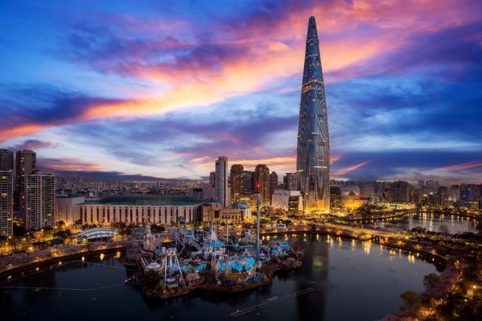 A panoramic view of a city skyline at sunset featuring a tall skyscraper and a waterfront amusement park.