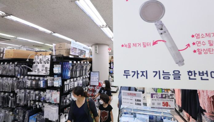 A view inside a store with shelves displaying various household items, including showerheads and filters, while shoppers browse the aisles. A promotional sign highlighting a multifunction water filter is visible in the foreground.