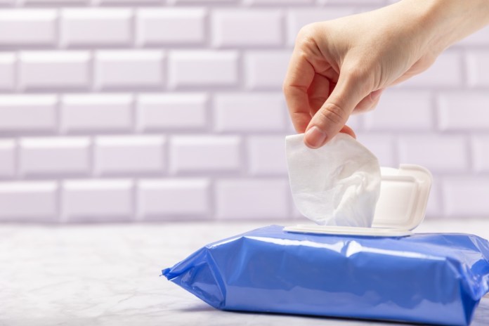 A hand pulling a wet wipe from a blue package, with a light-colored brick wall in the background.