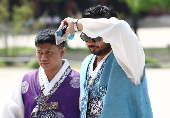 Two men standing outdoors wearing traditional Korean hanbok attire, with one shielding their eyes from the sun while the other looks ahead.