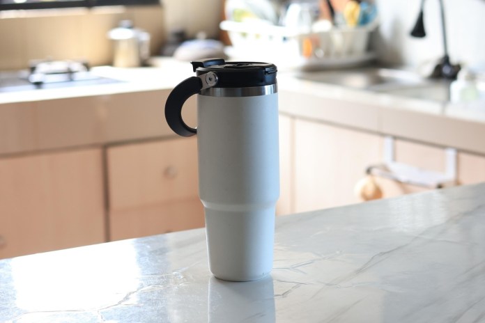 A white travel mug with a black lid and handle, placed on a marble countertop in a kitchen setting.