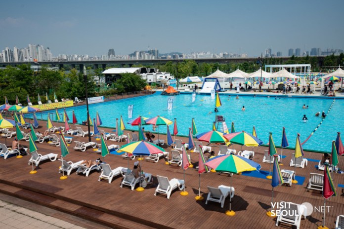 A vibrant outdoor swimming pool area featuring colorful umbrellas and sun loungers, with people enjoying the water on a sunny day. The city skyline is visible in the background.