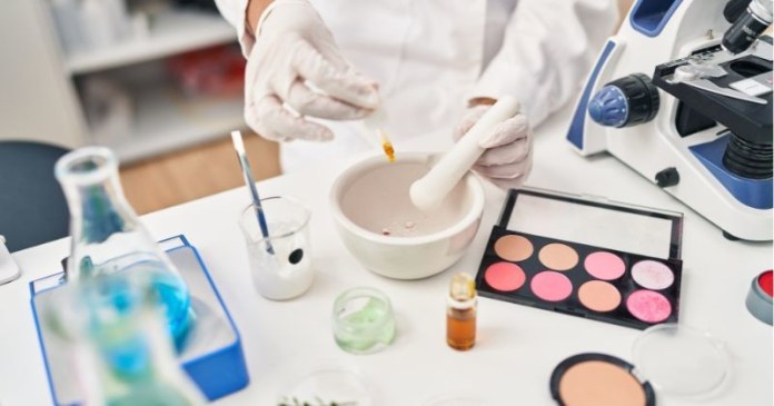 A person in a laboratory coat using a dropper to add liquid to a mortar and pestle, surrounded by various laboratory glassware and cosmetic products on a table.