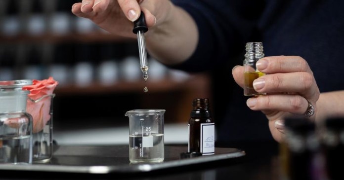 A person using a dropper to dispense liquid from a glass vial into a beaker on a lab tray, surrounded by various containers of liquids.