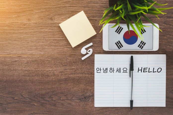 A flat lay image featuring a notepad with the words '안녕하세요' (Hello in Korean) and 'HELLO' written on it, a pen beside it, a yellow sticky note, a smartphone displaying the South Korean flag, and a small green plant on a wooden surface.