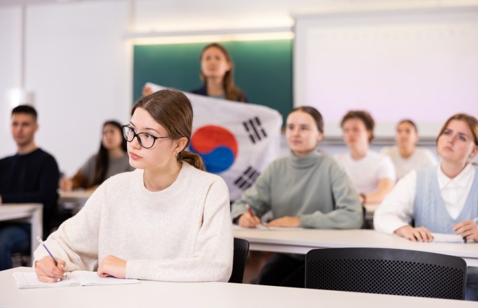 A classroom scene with diverse students listening attentively. A young woman in the foreground takes notes, while a classmate in the background holds up a South Korean flag.