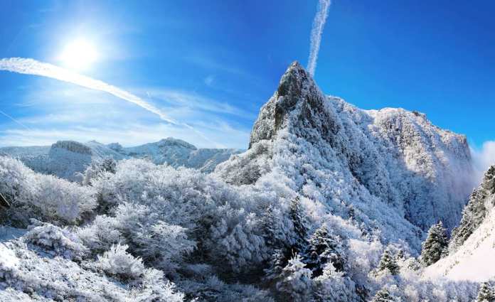 A panoramic view of snow-covered mountains and trees under a clear blue sky with the sun shining brightly.